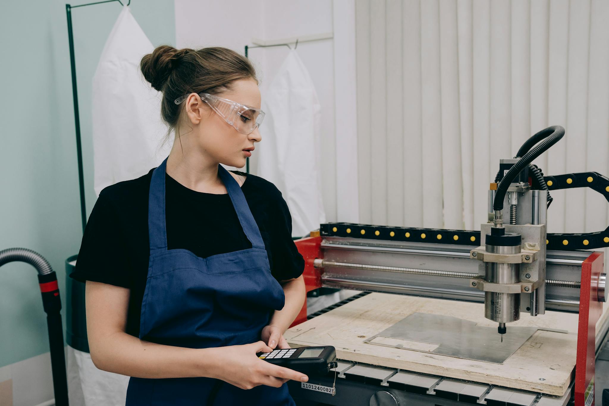 Woman in workshop using CNC machine, wearing goggles and apron, focused on industrial work.